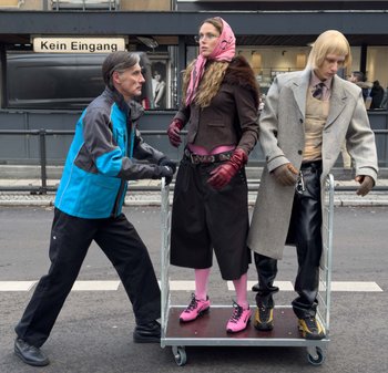 En mand iført en blå og grå jakke skubber en vogn med to påklædte dukker. Den ene har et pink tørklæde, en brun frakke og pink sko; den anden har en grå frakke og sorte bukser.