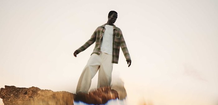 Mann trägt eine karierte Jacke und weite Hosen, steht auf felsigem Boden mit einer verschwommenen Reflexion und einem weichen beigefarbenen Himmel im Hintergrund.