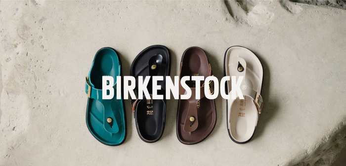 Vier einzelne Birkenstock-Sandalen in Türkis, Schwarz, Braun und Weiß nebeneinander auf einem strukturierten Betonboden angeordnet.