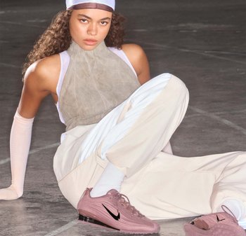 Young woman with curly hair in beige and white athletic outfit, seated on floor, wearing pink Nike sneakers and a white headband.