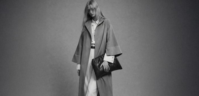 Femeie blondă purtând un palton lung și o ținută albă, ținând o geantă clutch mare, neagră, pe un fundal simplu.