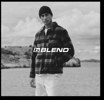 Zwart-witfoto van een man die een geruit jasje draagt met donkere en lichte tinten, een wit hemd en witte broek, tegen een achtergrond van water.