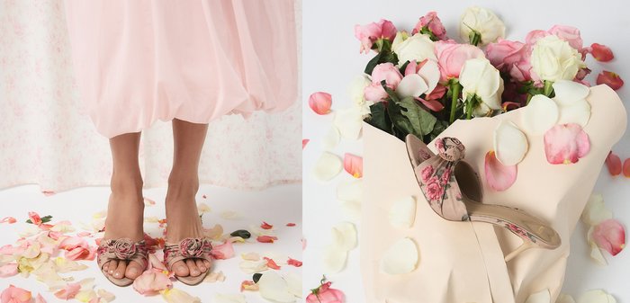 Füße in offenen Absätzen mit Blumenmuster und Rosenverzierung, stehend auf einem weißen Boden, umgeben von verstreuten rosa und weißen Blütenblättern.