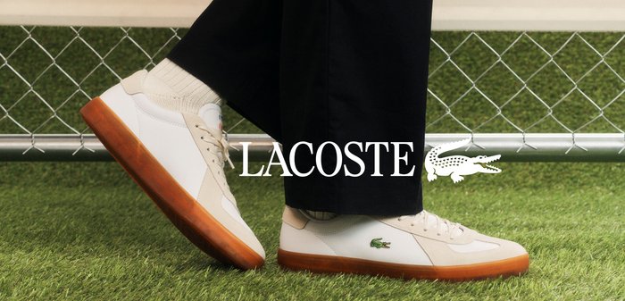 Person wearing white Lacoste sneakers with gum soles and white socks, standing on green artificial grass near a chain-link fence.