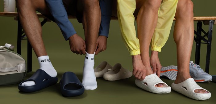 Zwei Personen sitzen und ziehen über Socken Schlupfschuhe an; eine trägt dunkelblaue Schuhe mit weißen Socken, die andere beigen Schuhe zu einem gelben Outfit.