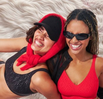 Due donne sorridono indossando un reggiseno sportivo nero con motivo, una sciarpa rossa e soffice e occhiali da sole. Lo sfondo presenta trame morbide e ondulate.