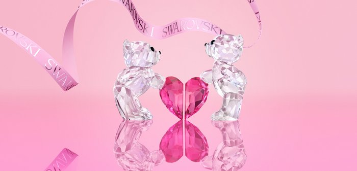 Twee transparante kristallen teddyberen die elkaar aankijken en elk een helft vasthouden van een roze kristallen hart met daarboven een Swarovski-lint.