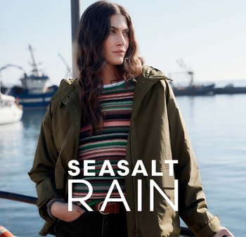 Olivově zelená bunda do deště s kapucí, s zipem. Pod ní je svetr s vícebarevnými pruhy a texturou. V pozadí voda a lodě.