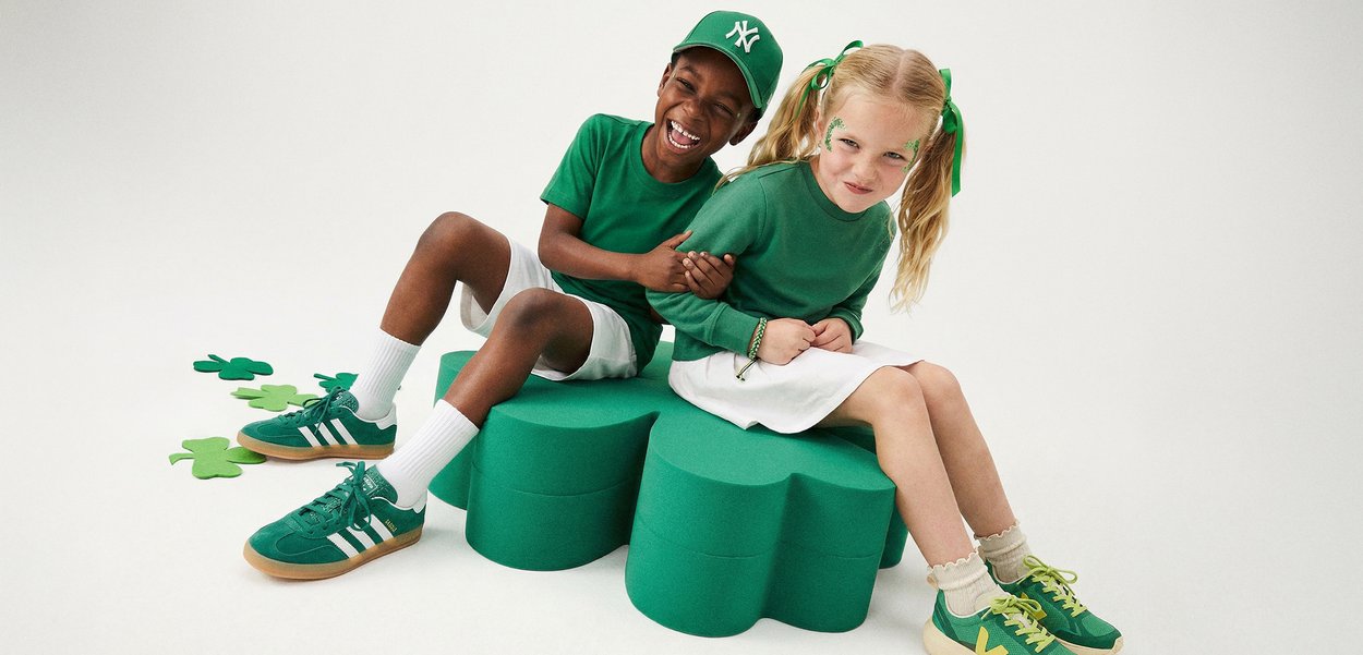 Two children dressed in green clothing and trainers sit on large green stools shaped like clovers, smiling and playing together.