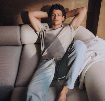 Ung man som lutar sig tillbaka på en ljusgrå soffa med händerna bakom huvudet, iklädd en beige t-shirt och ljusblå jeans i ett rum med träpanel.