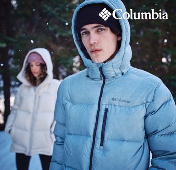 Junger Mann in blauer Columbia-Daunenjacke und schwarzem Beanie, Frau in weißer Jacke und pinker Mütze steht vor einem verschneiten Waldhintergrund.