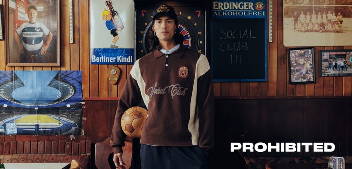 Young man in brown and beige sweater holding a soccer ball, standing in a room with vintage sports posters and a dartboard behind him.