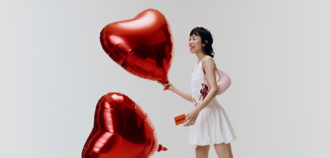Mlada ženska v beli obleki, ki drži dve rdeči srčni balon in oranžno Zalando kartico, se smehlja na svetlem enobarvnem ozadju.