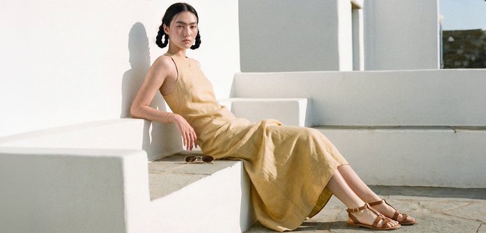 Femme aux cheveux tressés portant une robe beige et des sandales, assise sur des marches en béton blanc sous un soleil éclatant.