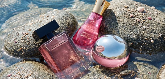 Trois flacons de parfum roses placés sur des rochers mouillés près d'une eau peu profonde avec de petites coquilles et un petit crabe à proximité.