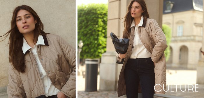 Jeune femme aux longs cheveux bruns porte une veste matelassée beige sur une chemise blanche, debout contre un mur clair en pierre avec une main dans la poche.