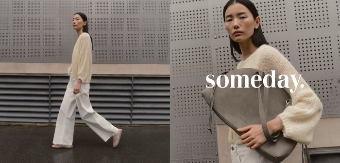 Femme aux longs cheveux foncés portant un pull crème tient un grand sac à main gris devant un mur en métal perforé avec le texte "un jour."