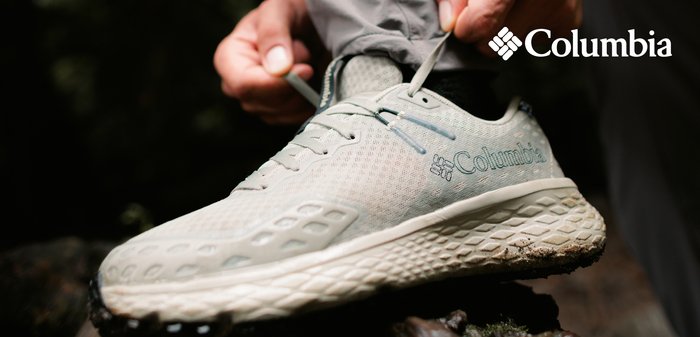 Person wearing gray Columbia hiking shoes ties laces while standing on a rock in an outdoor forest setting.