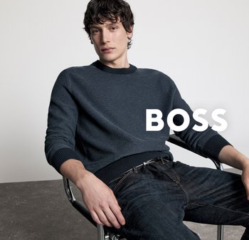 Junger Mann mit dunklem, welligem Haar, der einen marineblauen Pullover und dunkle Jeans trägt, entspannt auf einem Metallstuhl vor einem schlichten Hintergrund sitzt.