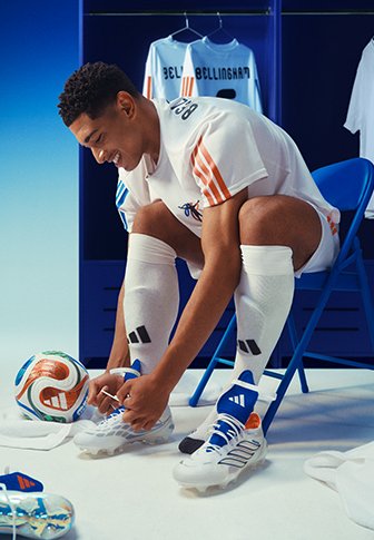 Jonge voetballer zittend op een blauwe stoel met het strikken van zijn voetbalschoenen, met een bal en shirts die op de achtergrond hangen.