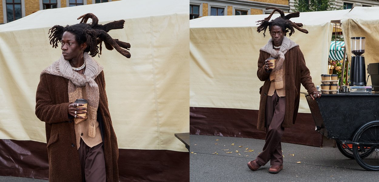 Manteau épais brun texturé sur un pull en laine beige et un pantalon brun foncé. Les accessoires incluent une écharpe épaisse et une tasse. Des dreadlocks sont soigneusement stylisées.