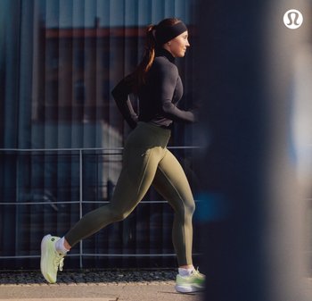 Zwart langemouwen top, olijfgroene high-waisted legging en lichtgroene hardloopschoenen. Op de achtergrond zijn verticale jaloezieën en een leuning te zien.