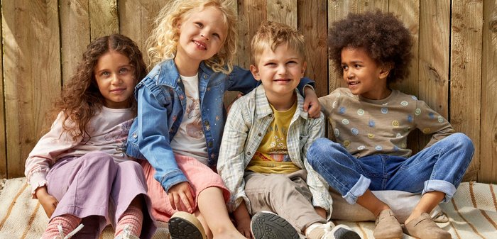 Vier Kinder posieren zusammen: eines trägt ein hellrosa, blumiges Langarmshirt, das zweite ein taupefarbenes Shirt mit bunten Punkten, das dritte ein gelbes T-Shirt mit Aufdruck und das vierte eine Jeansjacke mit floralem Stickerei über einem weißen Shirt. Sie tragen lässige Hosen und Röcke, mit Texturen von Baumwolle bis Denim.