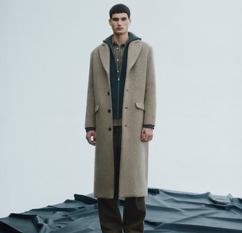 Beiger Wollmantel mit großen Revers, einreihiger Knopfleiste, darüber getragen eine marineblaue Jacke mit Reißverschluss und ein gemustertes Hemd. Braune Hose.