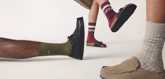 Vier Beine mit unterschiedlichen Socken, verschiedenen Schuhen und Sandalen, leicht angehoben vor einem schlichten Hintergrund.