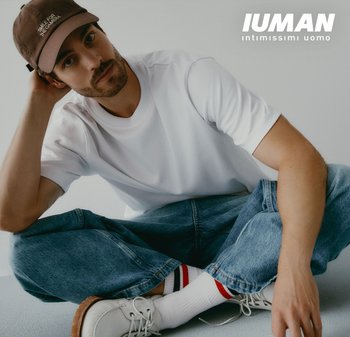 Mann in weissem T-Shirt, blauen Jeans, weissen Turnschuhen und braunem Hut, der im Schneidersitz gegen eine weisse Wand mit dem IUMAN Intimissimi Uomo Logo sitzt.