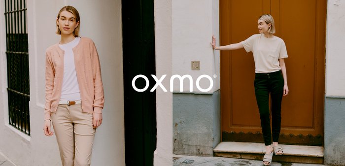 Frau mit kurzen blonden Haaren, trägt ein beigefarbenes Hemd und schwarze Hose, steht vor einer braunen Tür, eine Hand berührt die weiße Wand.