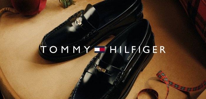 Schwarze Lackleder-Slipper mit einem dekorativen Riemen und Logo-Beschlägen, vor einem beige Hintergrund mit einem karierten Akzent.