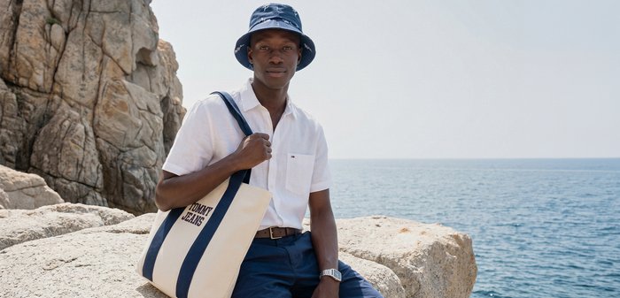 Νέος άνδρας που φοράει καπέλο τύπου bucket και λευκό πουκάμισο κάθεται σε βραχώδη ακτή κρατώντας μια τσάντα από καμβά δίπλα σε ήρεμη θάλασσα κάτω από καθαρό ουρανό.