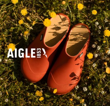 Sabots en caoutchouc rouge avec une texture lisse, bout arrondi et semelle en liège, posés sur de l'herbe entourée de fleurs jaunes. Logo visible.