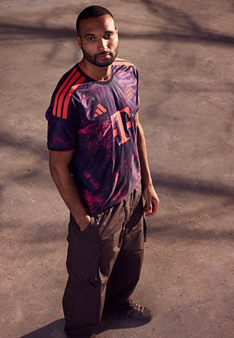 Mann mit kurzen Haaren und Bart, der ein dunkelviolettes und rotes Sporttrikot sowie braune Cargohosen trägt, steht auf dem Bürgersteig.