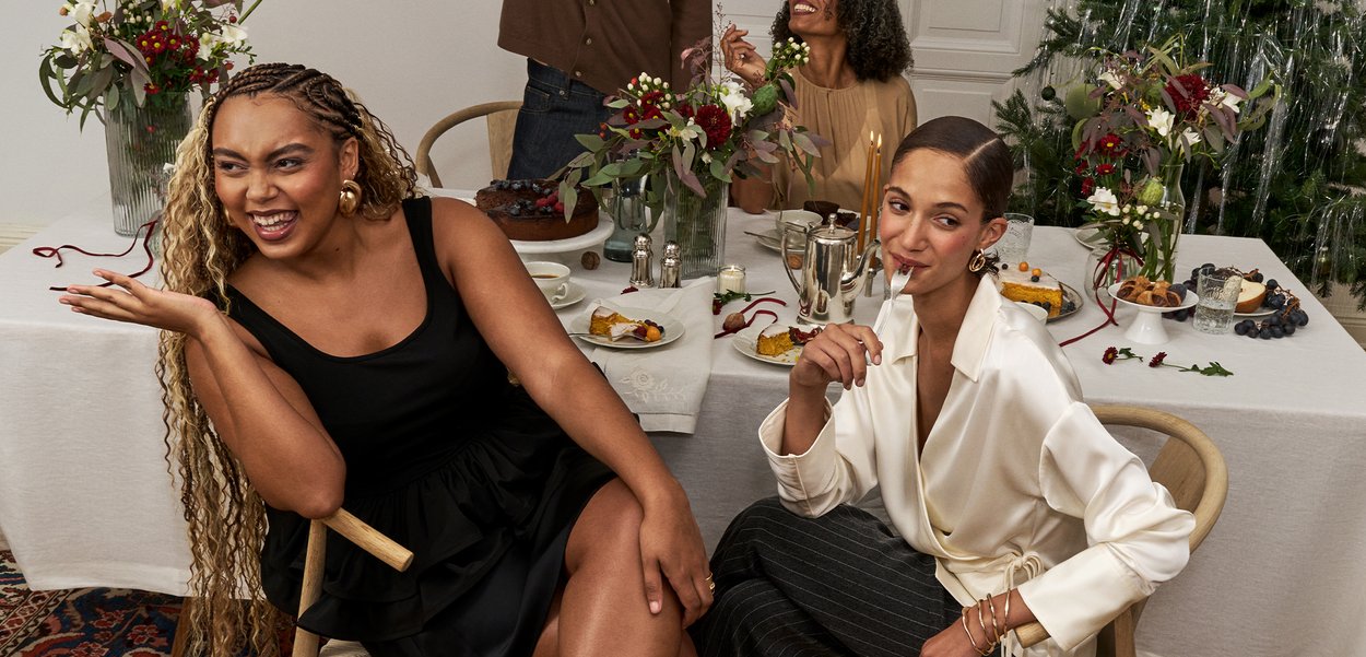 Twee vrouwen zitten aan een eettafel die is gedekt voor een maaltijd. De ene draagt een zwarte jurk, de andere een witte blouse en gestreepte broek; beiden hebben accessoires.