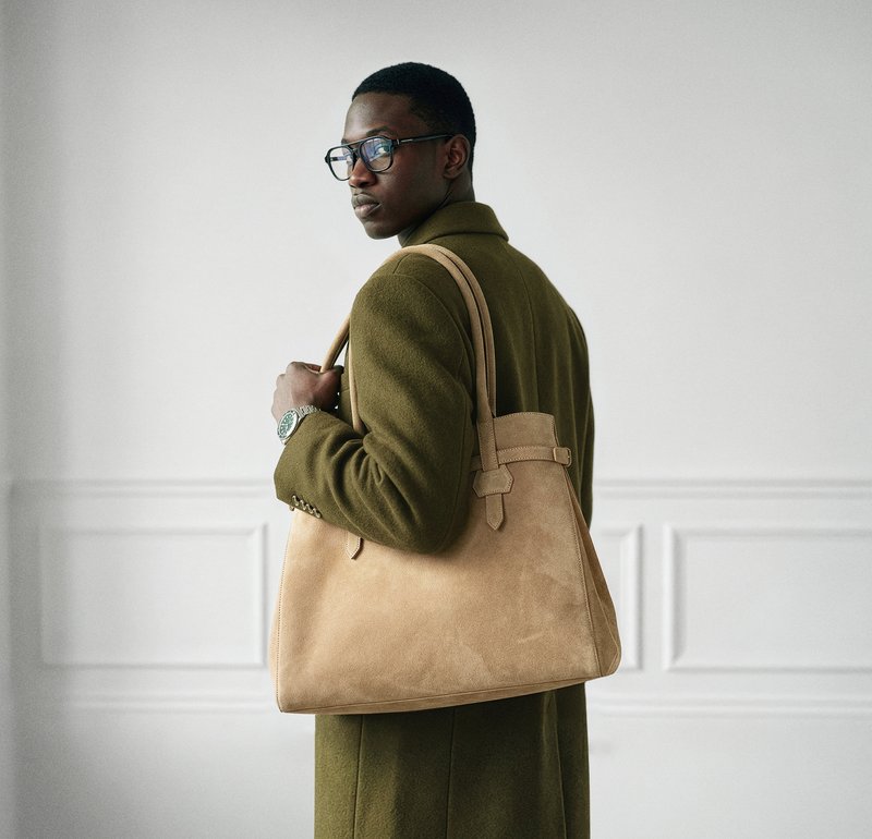 Vīrietis ar brillēm un olīvu zaļas krāsas mēteļu, pie pleca nēsājot lielu tanādas somu, stāv minimālistiskā baltā istabā.