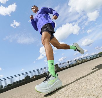 Hardloper die een blauwe jas, zwarte korte broek en groene sokken met witte hardloopschoenen draagt, joggend buiten op een zonnige dag.