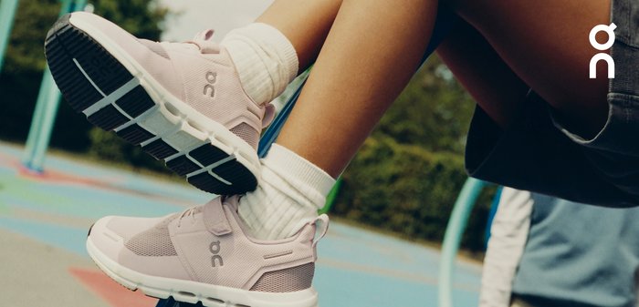 Witte sportschoenen met een bovenwerk van mesh, zwarte veters en een gevoerde zool met uitgesproken rubberen accenten. Gecombineerd met lichtgrijze broek.