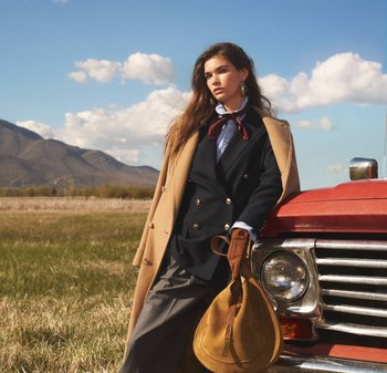 Modello che indossa un blazer doppiopetto blu navy, una camicia azzurro chiaro e guanti in pelle marroni. Cappotto beige appoggiato sopra. Tiene una borsa in suede color tan.