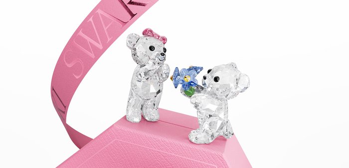 Deux ours en cristal clair sur une boîte rose, l'un tenant une fleur bleue, entourés d'un ruban rose à la marque Swarovski.