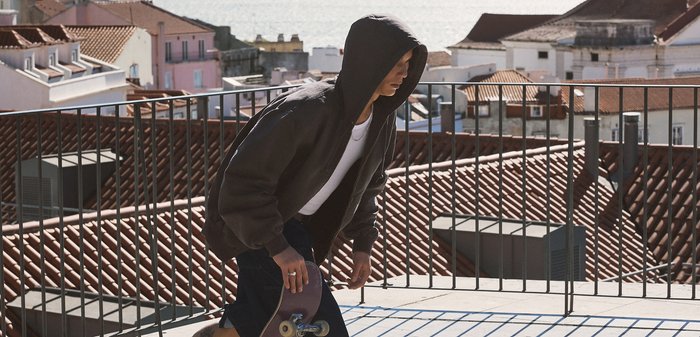 Νέος άνθρωπος με μαύρο φούτερ με κουκούλα καθισμένος σκυφτός κρατά ένα σκέιτμπορντ σε μια στέγη με τερακότα πλακίδια και κτίρια στην παραλία στο βάθος.