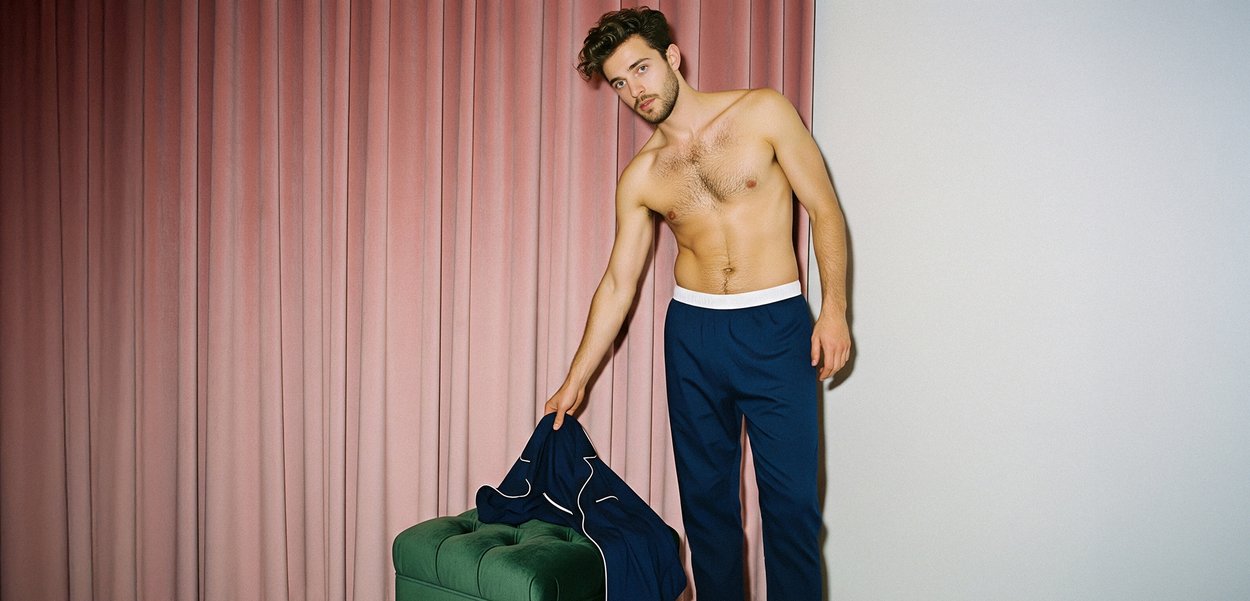 Παντελόνι πιτζάμας σε ναυτικό μπλε με λευκή ζώνη, συνδυασμένο με ναυτικό μπλε σακάκι. Το μοντέλο στέκεται κοντά σε οθωμανικό κάθισμα με πράσινες λεπτομέρειες και μαλακό ροζ φόντο.