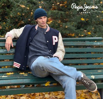 Giacca varsity blu navy e panna con logo "P", polsini a coste, maglione grigio lavorato a maglia, jeans azzurri e sneakers scure, seduto su una panchina verde.