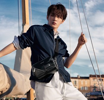 Junger Mann mit einer marineblauen Jacke und einer Umhängetasche, der ein Segelbootseil hält, mit einer Marina und blauem Himmel im Hintergrund.