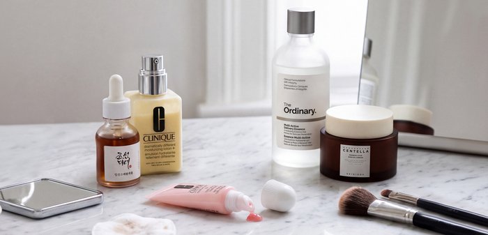 Huidverzorgingsproducten inclusief serums, moisturizer, essence, crème, make-upborstels, wattenschijfjes en een kleine spiegel op een marmeren oppervlak.