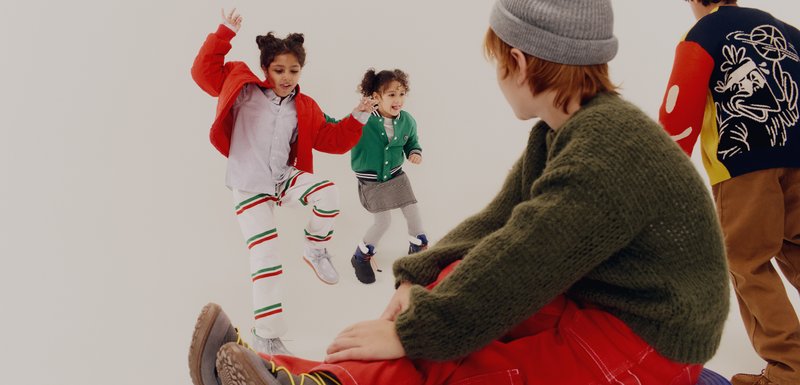Deux enfants en tenues colorées jouent : l'un porte une veste bomber rouge et un pantalon blanc rayé ; l'autre porte un cardigan vert et une jupe à carreaux.