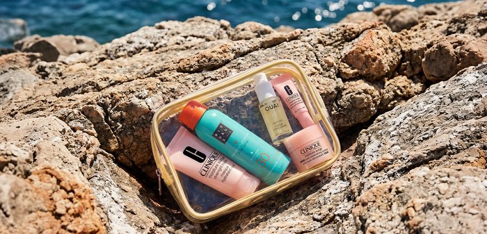Trousse de toilette transparente avec des flacons de crème solaire, soins de la peau et huile pour les cheveux, posée sur une rive rocheuse ensoleillée au bord de la mer.