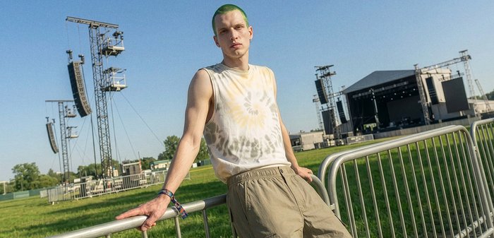 Jeune personne aux cheveux verts, tee-shirt tie-dye sans manches et pantalon beige appuyée sur des barrières métalliques sur une scène de concert en plein air en journée.