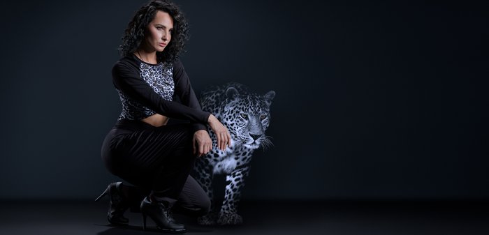 Zwart cropped top met luipaardprint, zwarte broek en zwarte hoge hakken. Er verschijnt een gedeeltelijk doorschijnende luipaard ernaast. Donkere achtergrond.